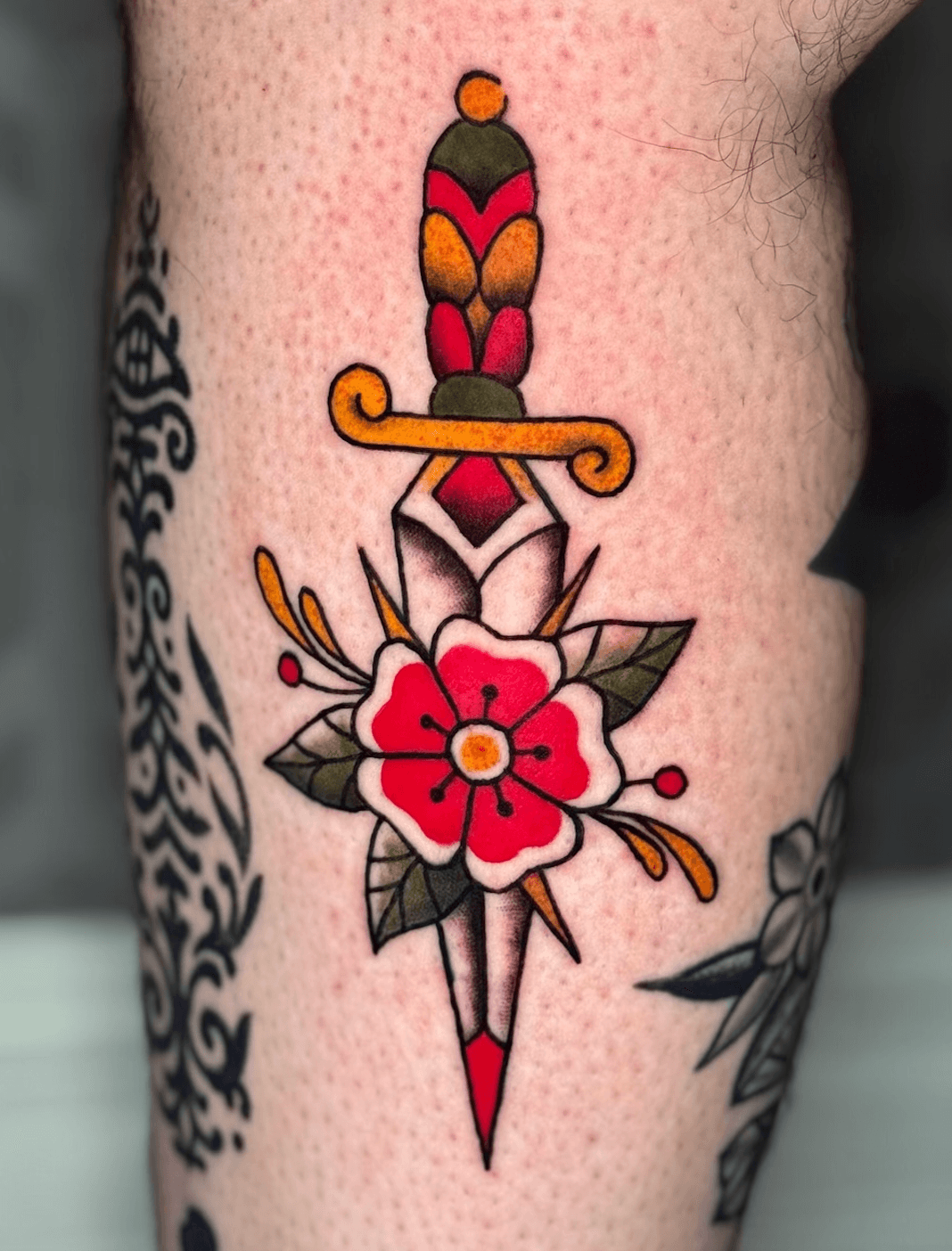 Colour traditional-style dagger tattoo with a red flower and green leaves