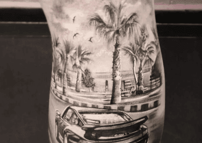 Detailed black-and-gray tattoo shows a car with an 'EST-1979' license plate, palm trees, and birds in flight.