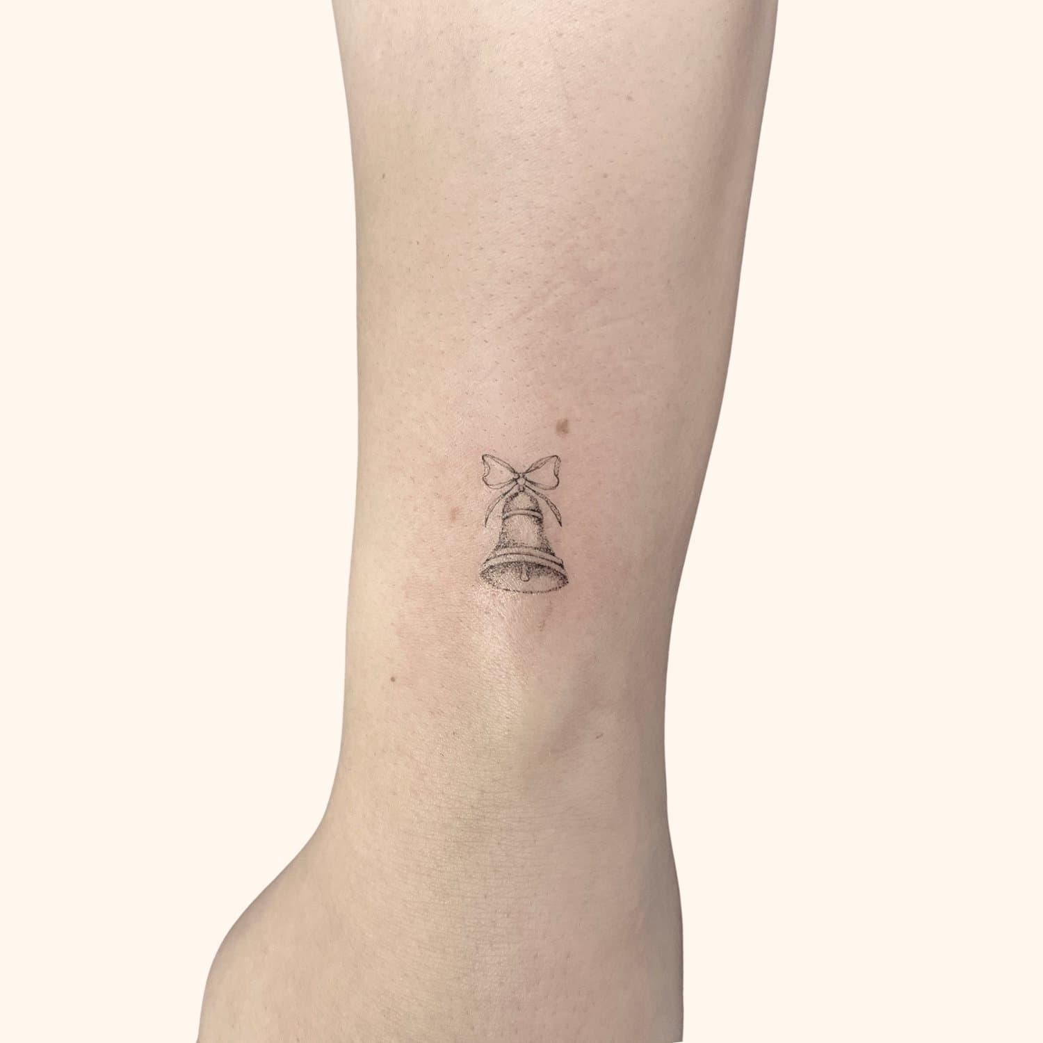 A small tattoo of a school bell with a ribbon bow on top, located on a person's inner ankle.