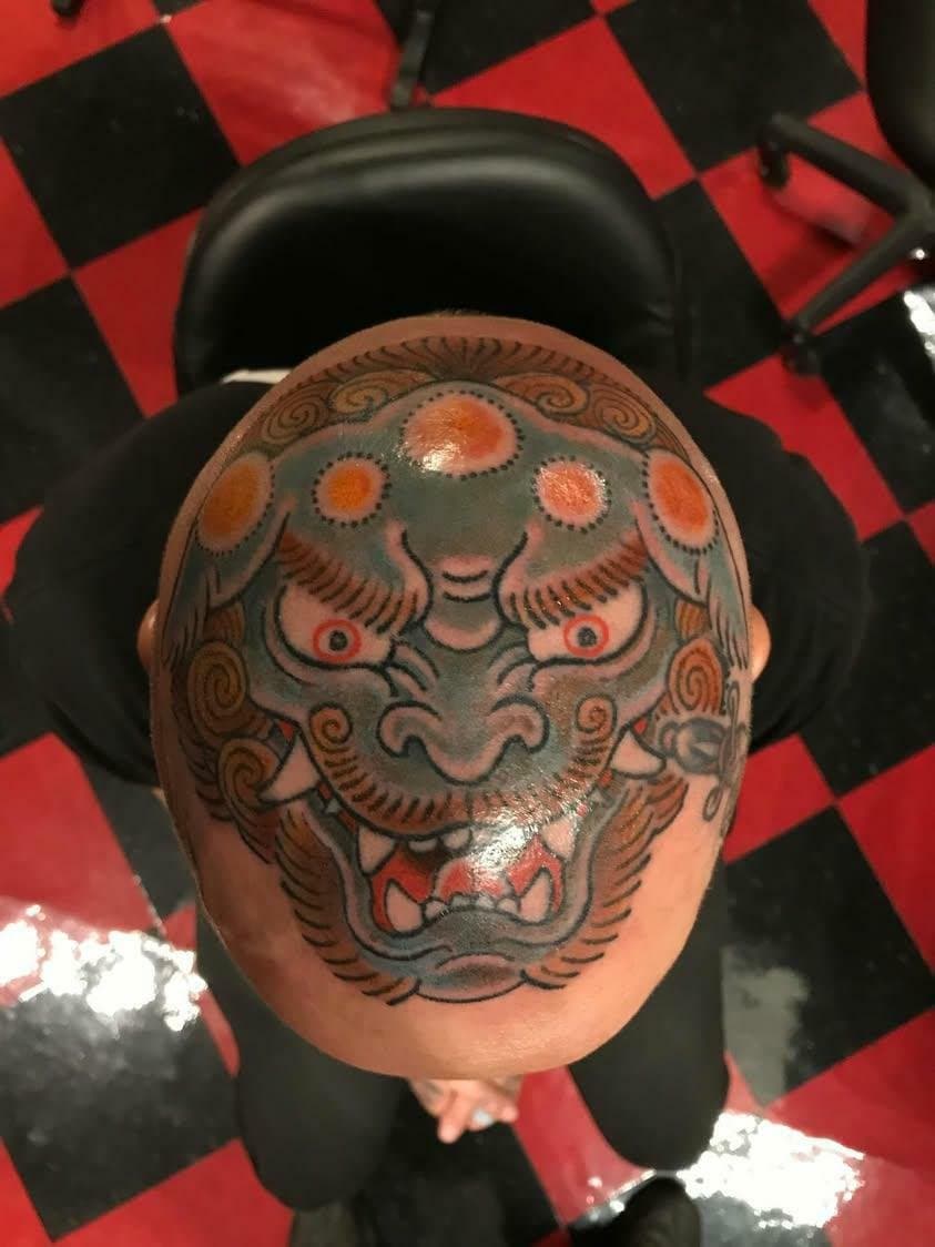 Tattoo of a stylistic dragon face on a person's bald head, with a red and black checkered floor in the background.