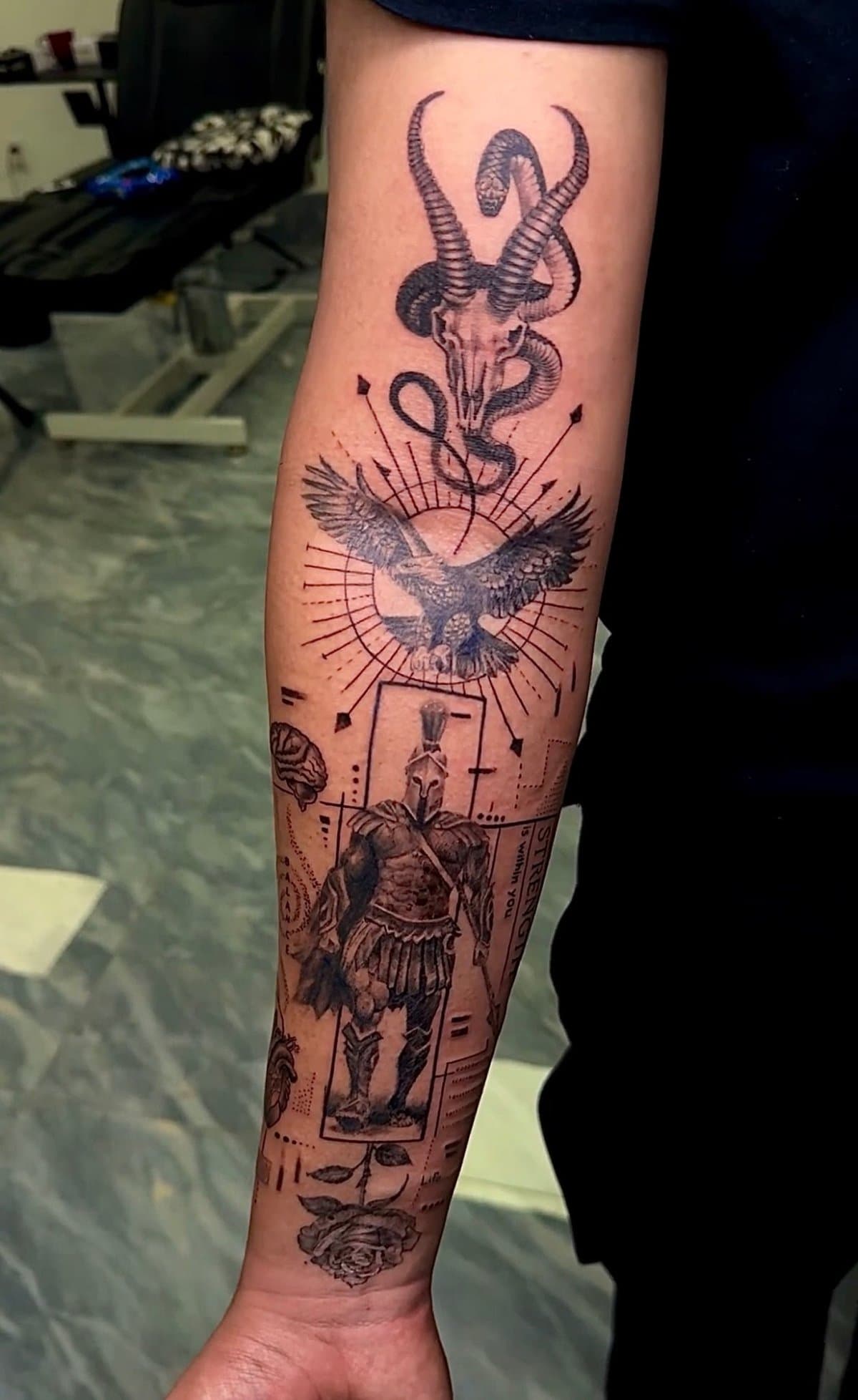Arm featuring a complex black ink tattoo with a skull, snake, eagle, gladiator, and rose.