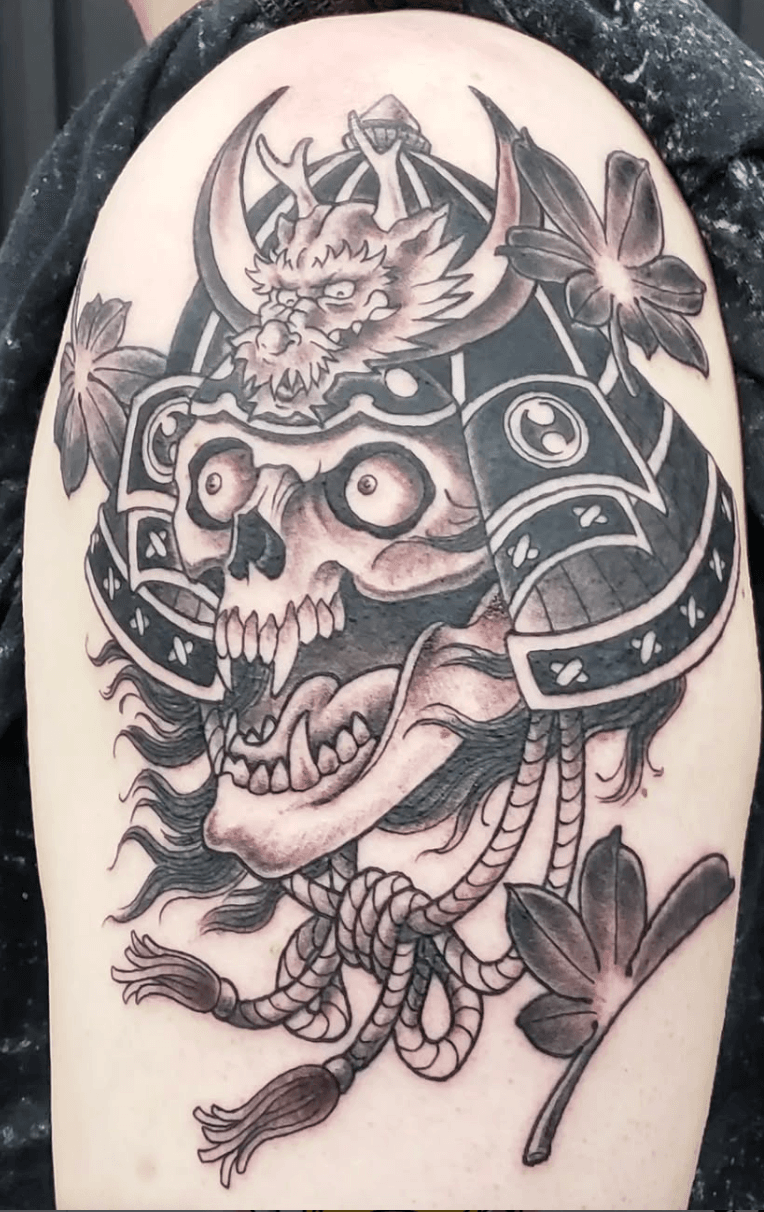 Tattoo of a fearsome skull with monstrous features, horns, and a devilish face, adorned with flowers and leaves, with ropes hanging below.