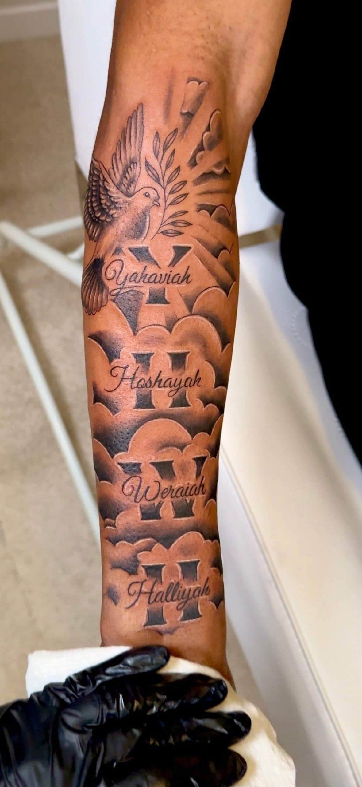 Tattoo of a dove, clouds, and names Yakoviah, Hoshayah, Weraiah, and Halliyah on a dark-skinned arm.