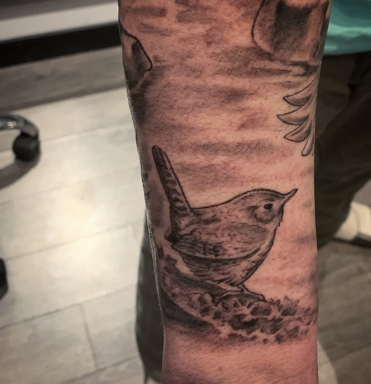 Black ink tattoo of a small bird, possibly a sparrow, perched on a branch.
