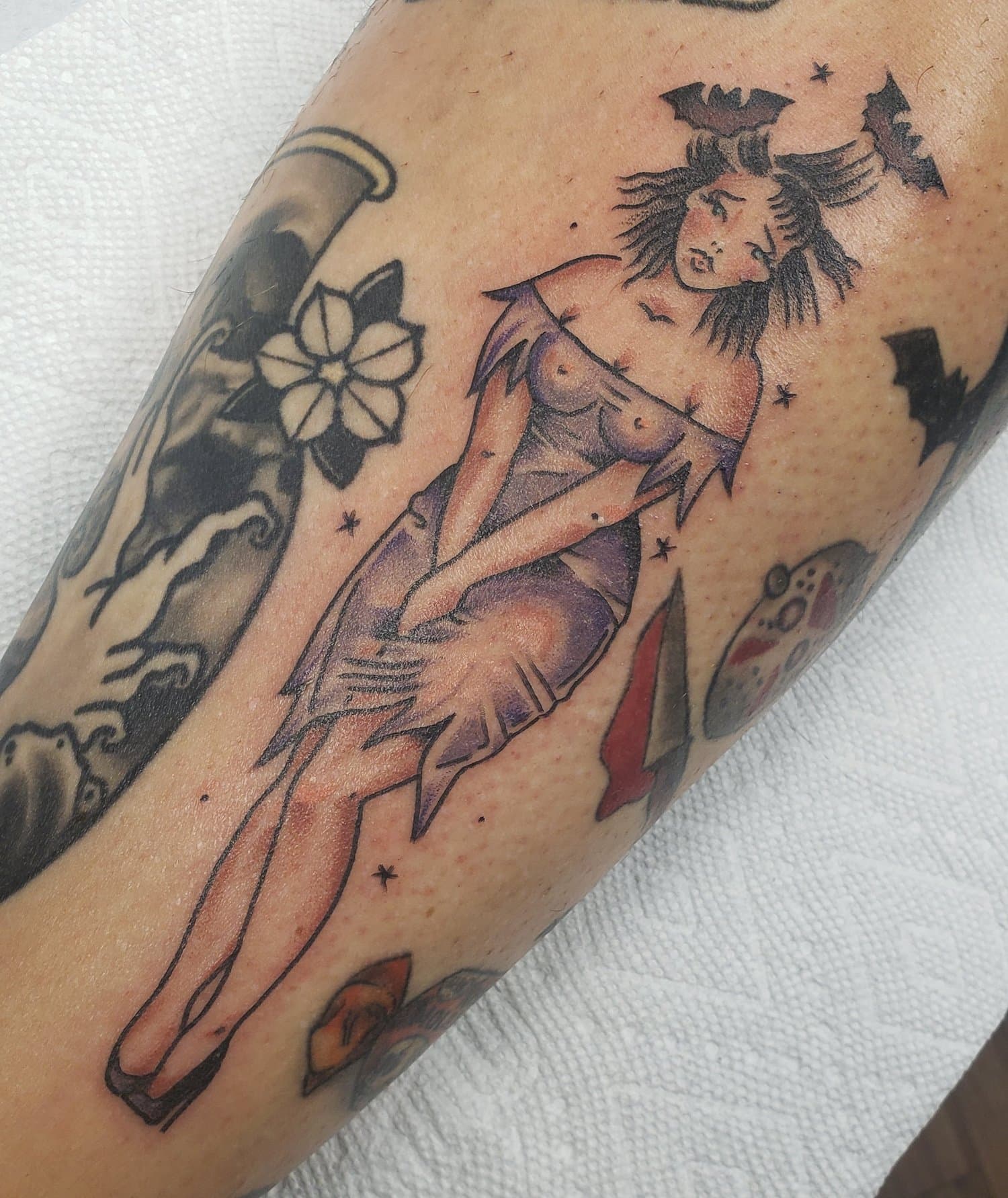 Tattoo of a woman with bat wings, sitting in a distressed pose, surrounded by stars, a flower, a paint palette, a lipstick, and a long object, with a mix of bla