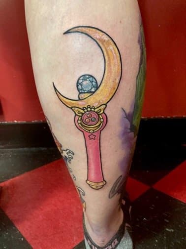 Tattoo of a pink sword with a crescent moon and a blue geometric shape on a person's leg.