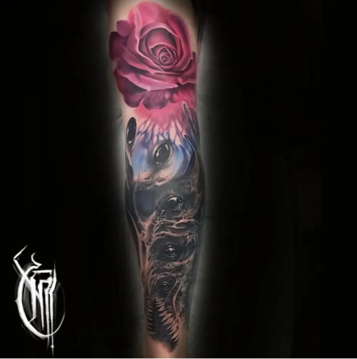 Forearm tattoo combining a pink and purple rose with a black and grey skull below it, carrying the artist's monogram watermark
