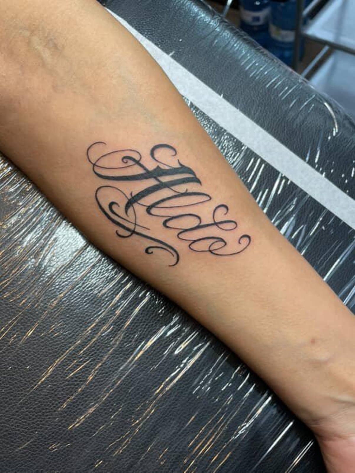 Arm displays black calligraphy tattoo with the name "Alda" in cursive script.