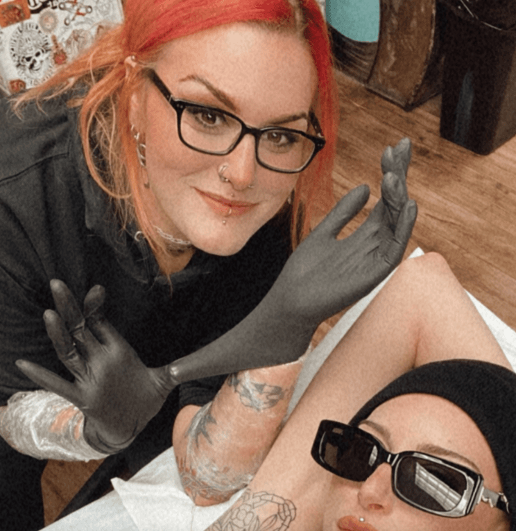 A woman with red hair, glasses, and tattoos is getting a tattoo on her arm by a tattoo artist. The artist is wearing black gloves and has tattoos on her arm.