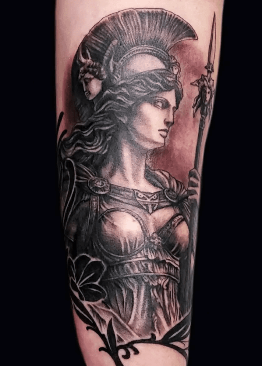 Black and gray tattoo of a Greek warrior woman wearing a helmet and armor, holding a spear, with decorative floral elements around her.
