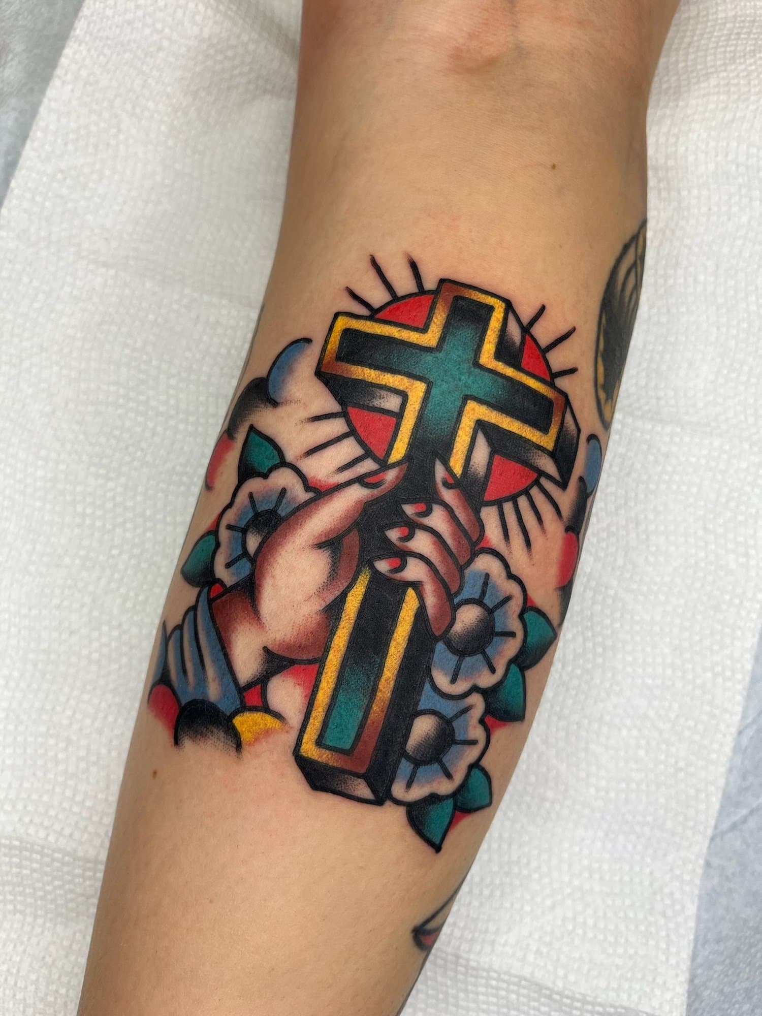 A colorful tattoo of a hand holding a cross, surrounded by flowers and a red sun with radiating lines.
