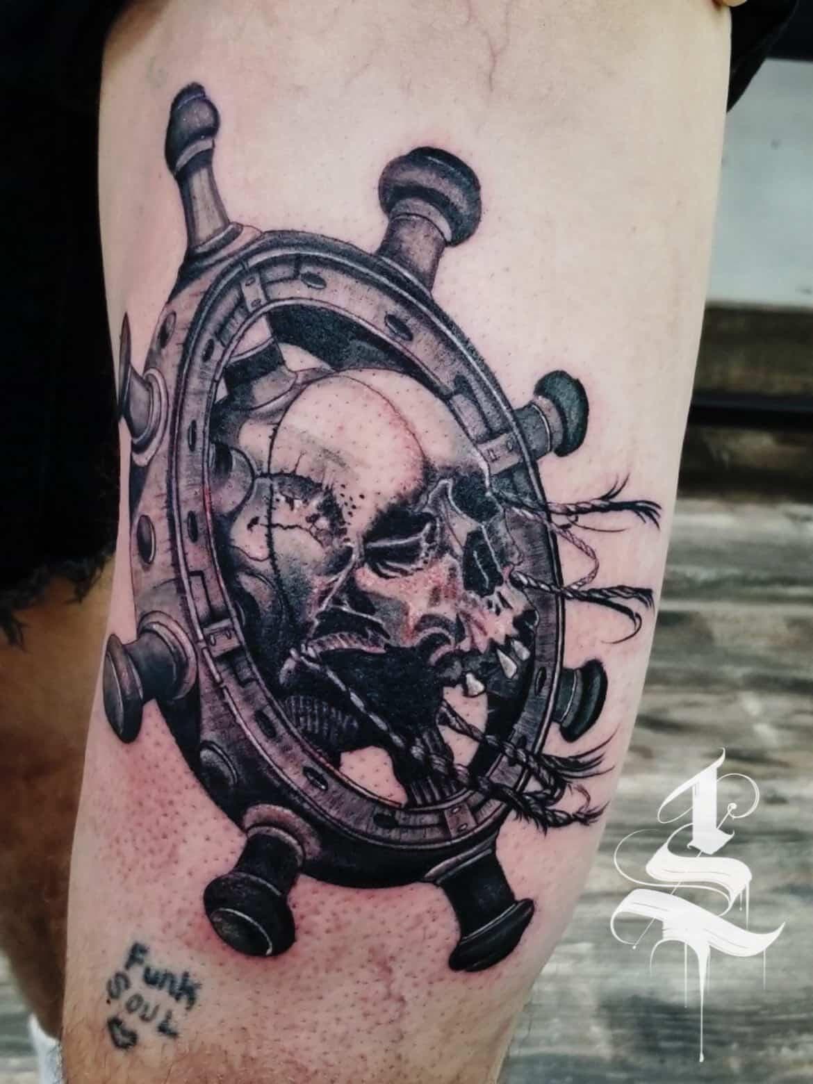 Thigh tattoo displays a ship's wheel enclosing a skull with rope accents.