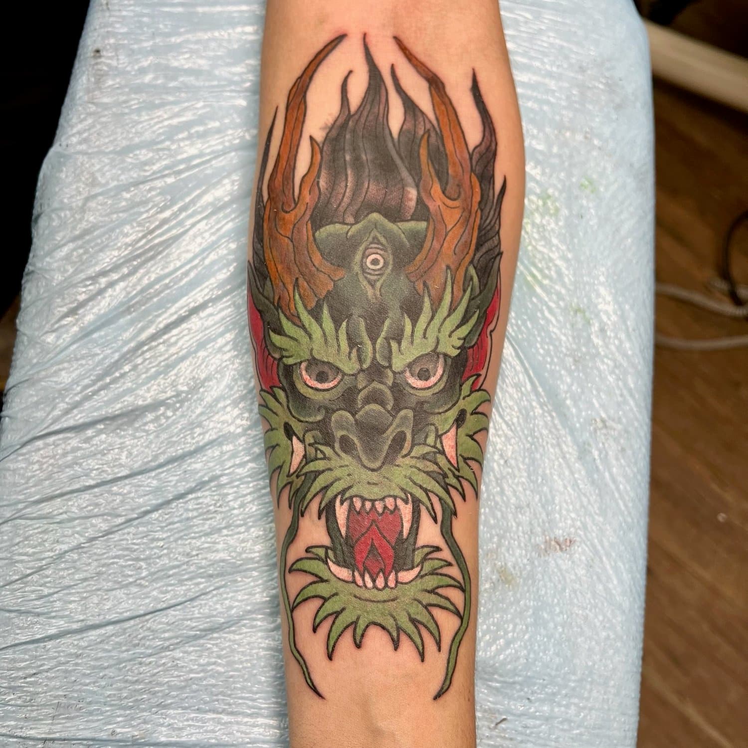 Colorful tattoo of a fierce dragon with green scales, sharp teeth, pink eyes, and red tongue on a person's arm. It is also a cover up tattoo.