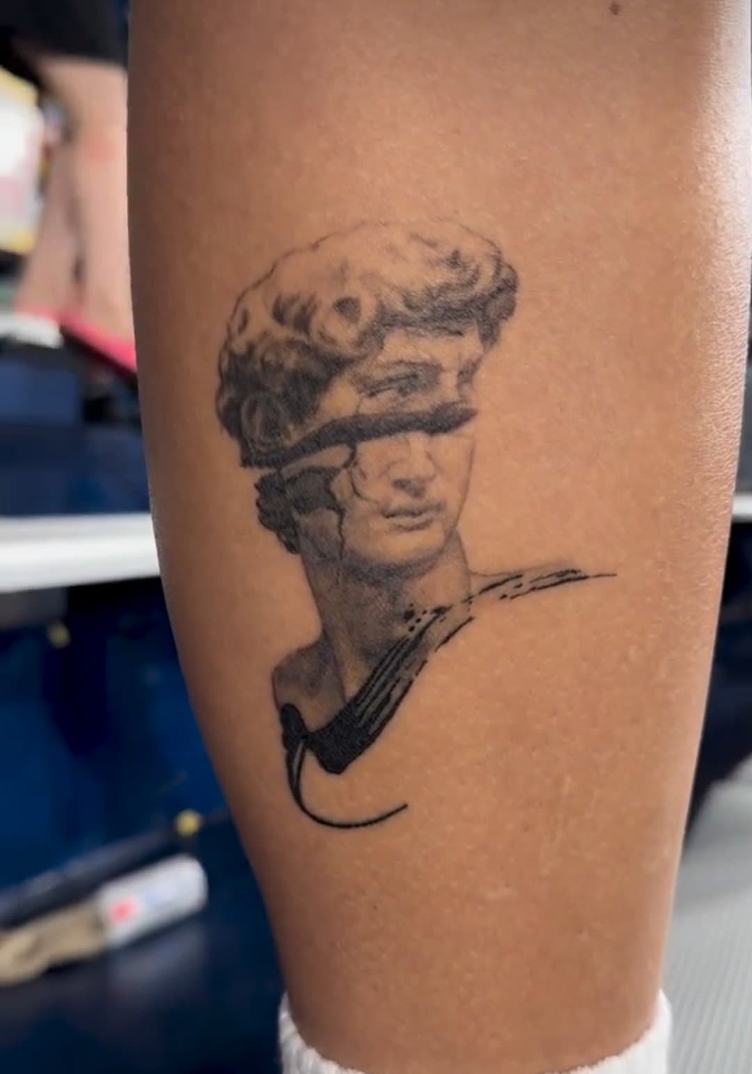 A tattoo of a bust with a crack across the face, on a person's arm.