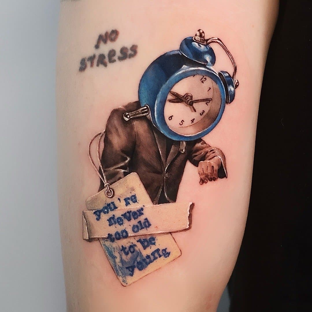 Tattoo of a man with a blue alarm clock as a head, wearing a suit. The clock shows time as 2:15. The man is holding a sign that says, "You'd be never too old to