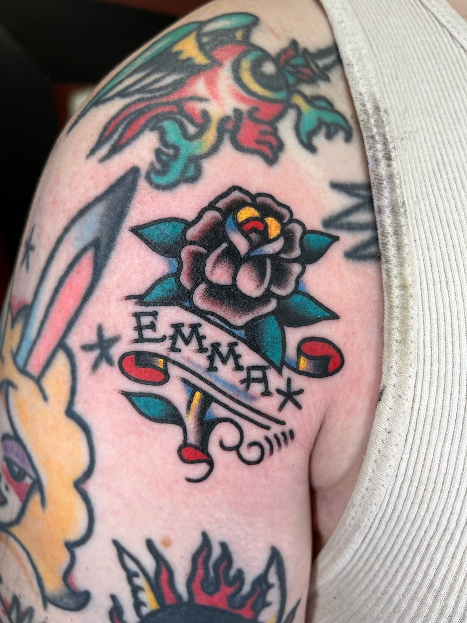 Tattoo of a rose with the word "EMMA" beneath it on upper arm.