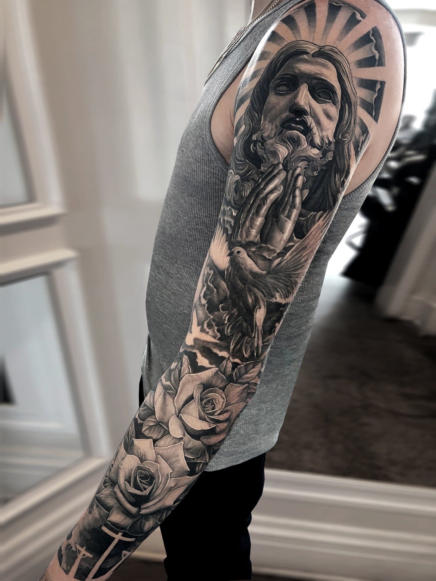 A detailed black and gray tattoo sleeve featuring a depiction of Jesus Christ with praying hands, a dove, and roses.