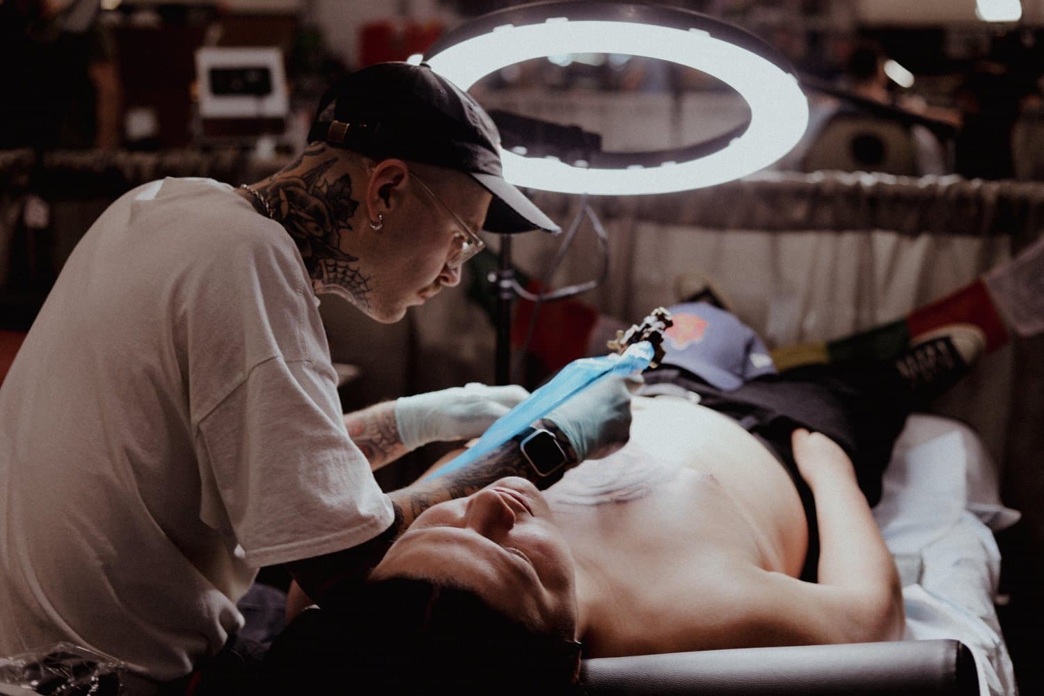 Tattoo artist working on a tattoo of a guitar on a client's arm, lying on a bed in a tattoo studio with a lamp overhead.
