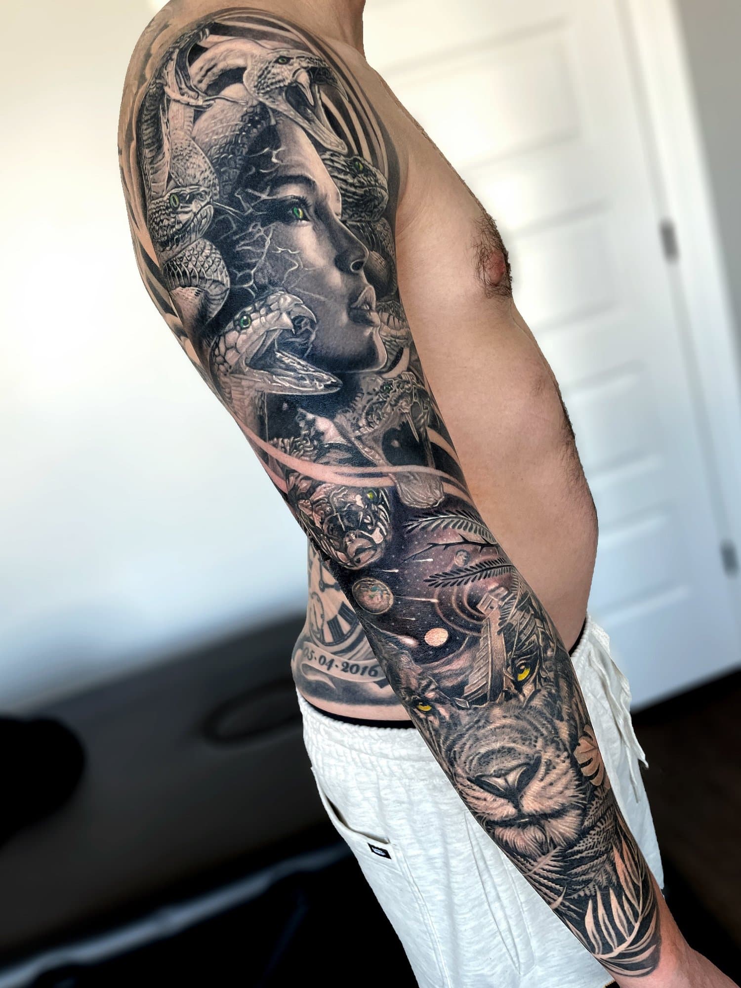 A man's arm tattoo sleeve featuring a woman's face, a lion's face, and various nature and fantasy elements in black and gray ink.