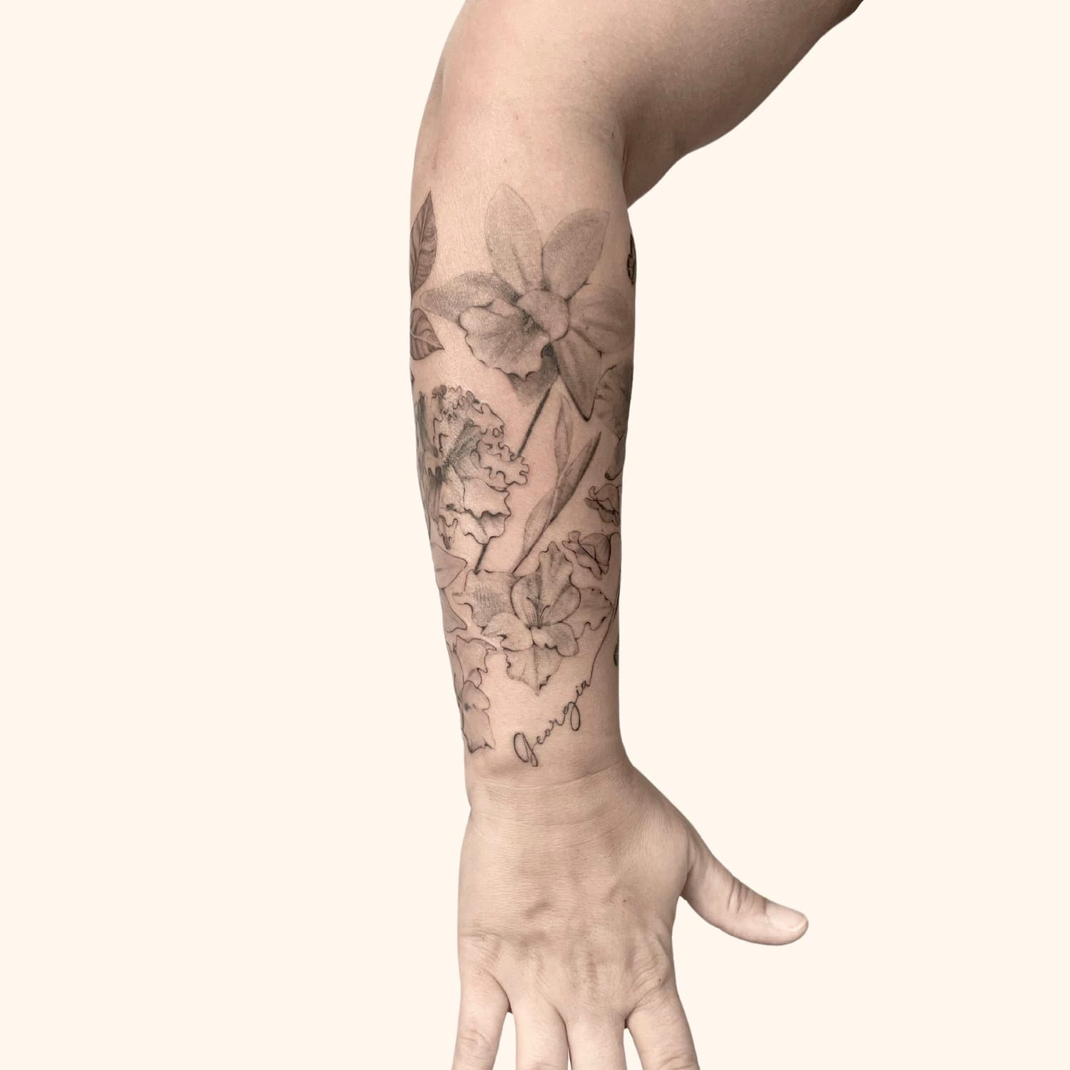 A person's arm with a detailed black and gray floral tattoo, including daffodils and leaves, with the name 'Jeorgia' tattooed at the bottom.
