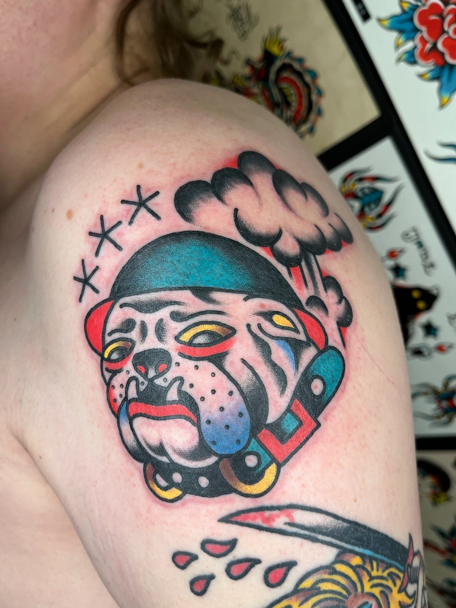 Traditional style tattoo of a bulldog wearing a helmet, with a smoking pipe, and drops of blood or paint, on a person's upper arm.