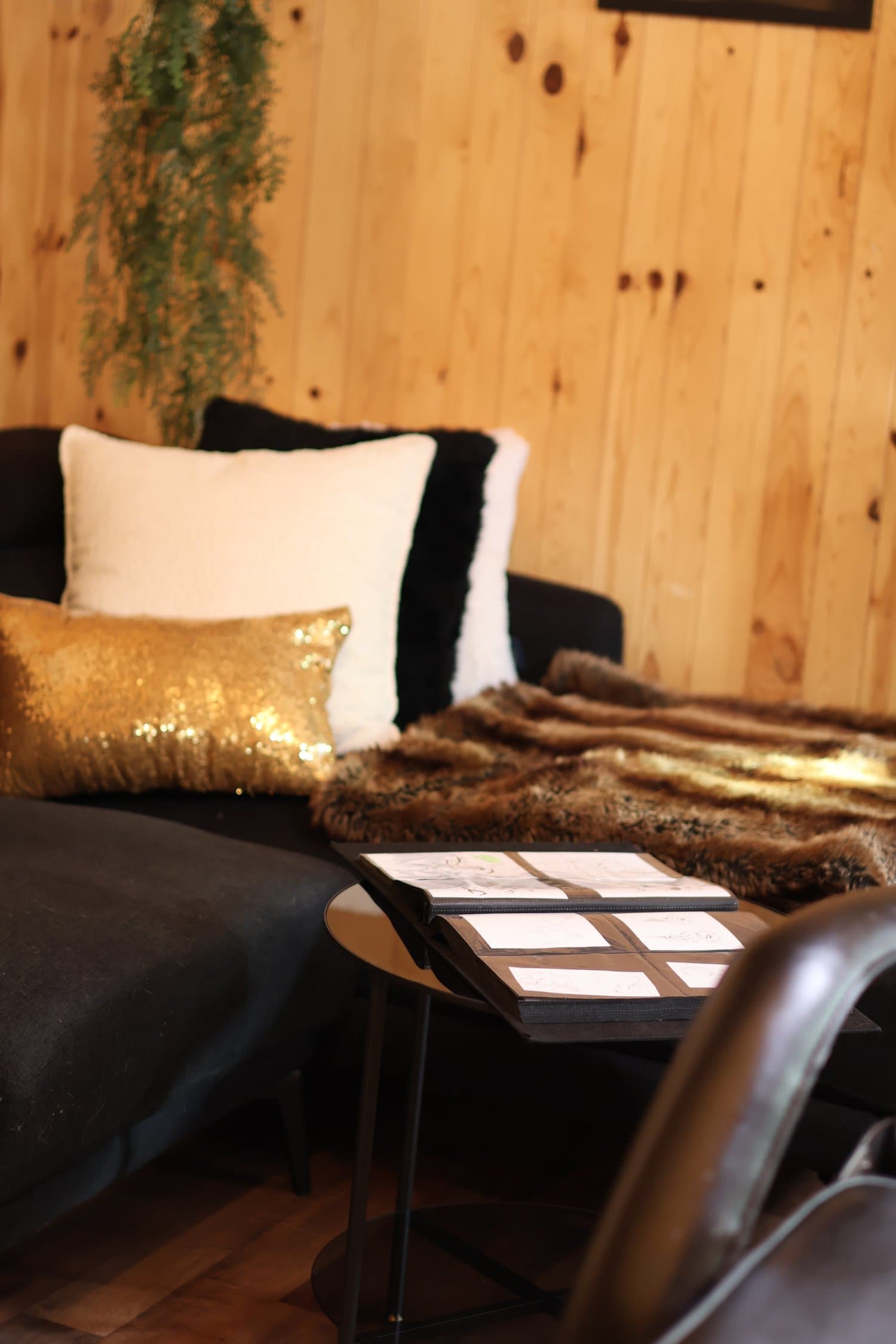 Un canapé noir avec plusieurs coussins, dont un doré à sequins, un blanc, et un noir, avec une couverture en fourrure, devant un mur en bois. Un petit meuble no