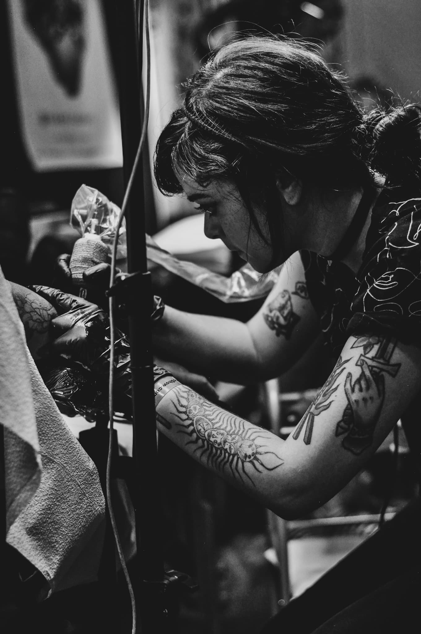 Black and white first-party photograph of a tattooed artist with her hair up working on a piece in progress, published in the Dallas Conte section of the studio roster page