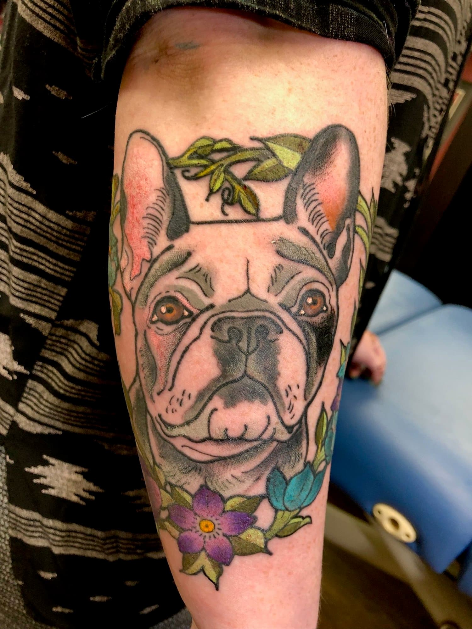 Tattoo of a French Bulldog's face with surrounding flowers and green leaves on a person's arm.