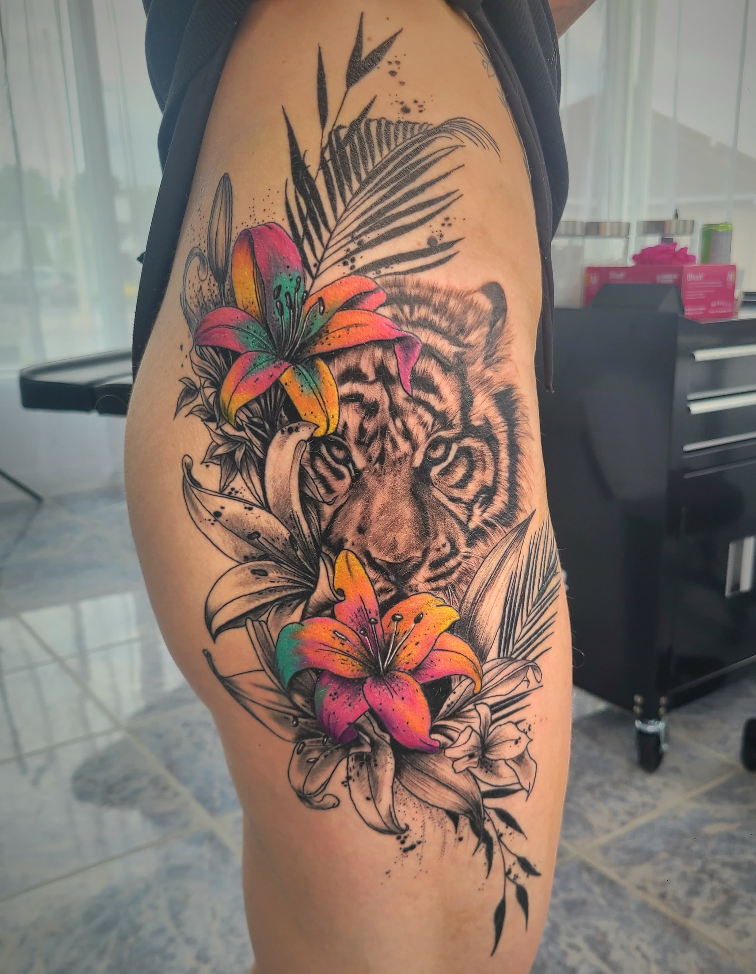 Thigh tattoo of a tiger face framed by tropical leaves in black and grey with orange and pink lilies in colour