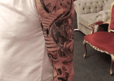 Detailed forearm tattoo displays a clock, stairs, a figure, and a woman angel, showcasing realistic black-and-gray inking.