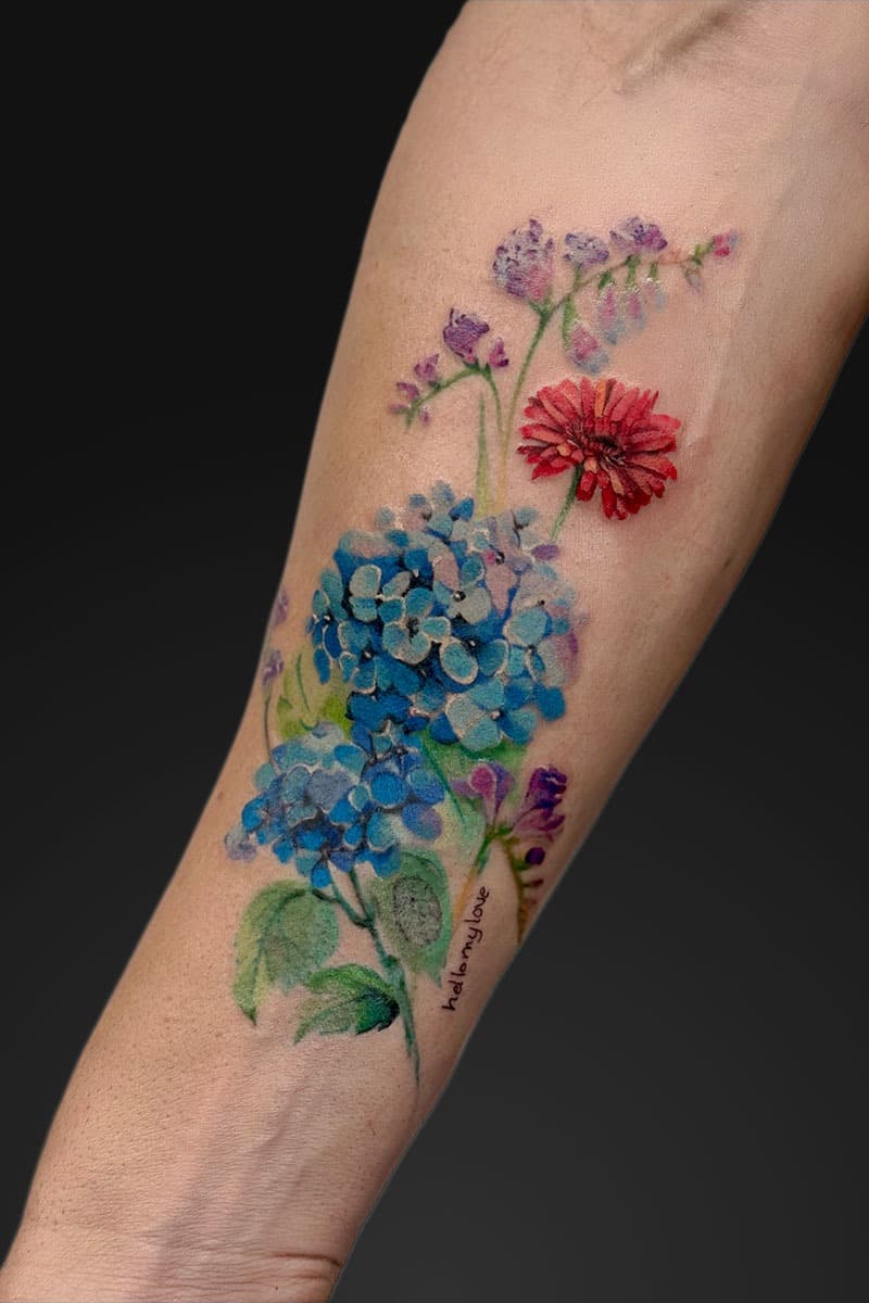 Watercolor-style forearm tattoo of blue hydrangea clusters with purple blossoms and a red bloom, green stems, and small script text along the stem.
