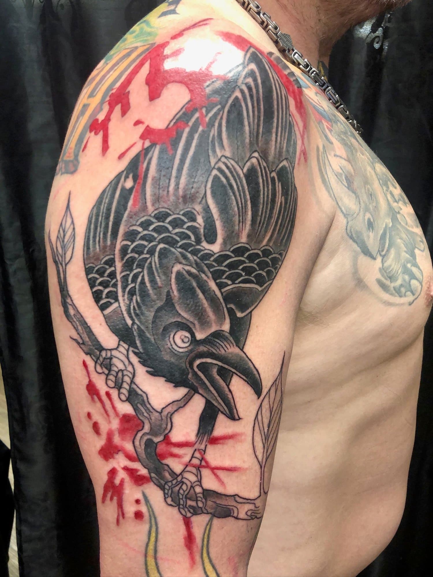 Tattoo of a raven perched on a branch with ink splatters and red accents on the person's upper arm.