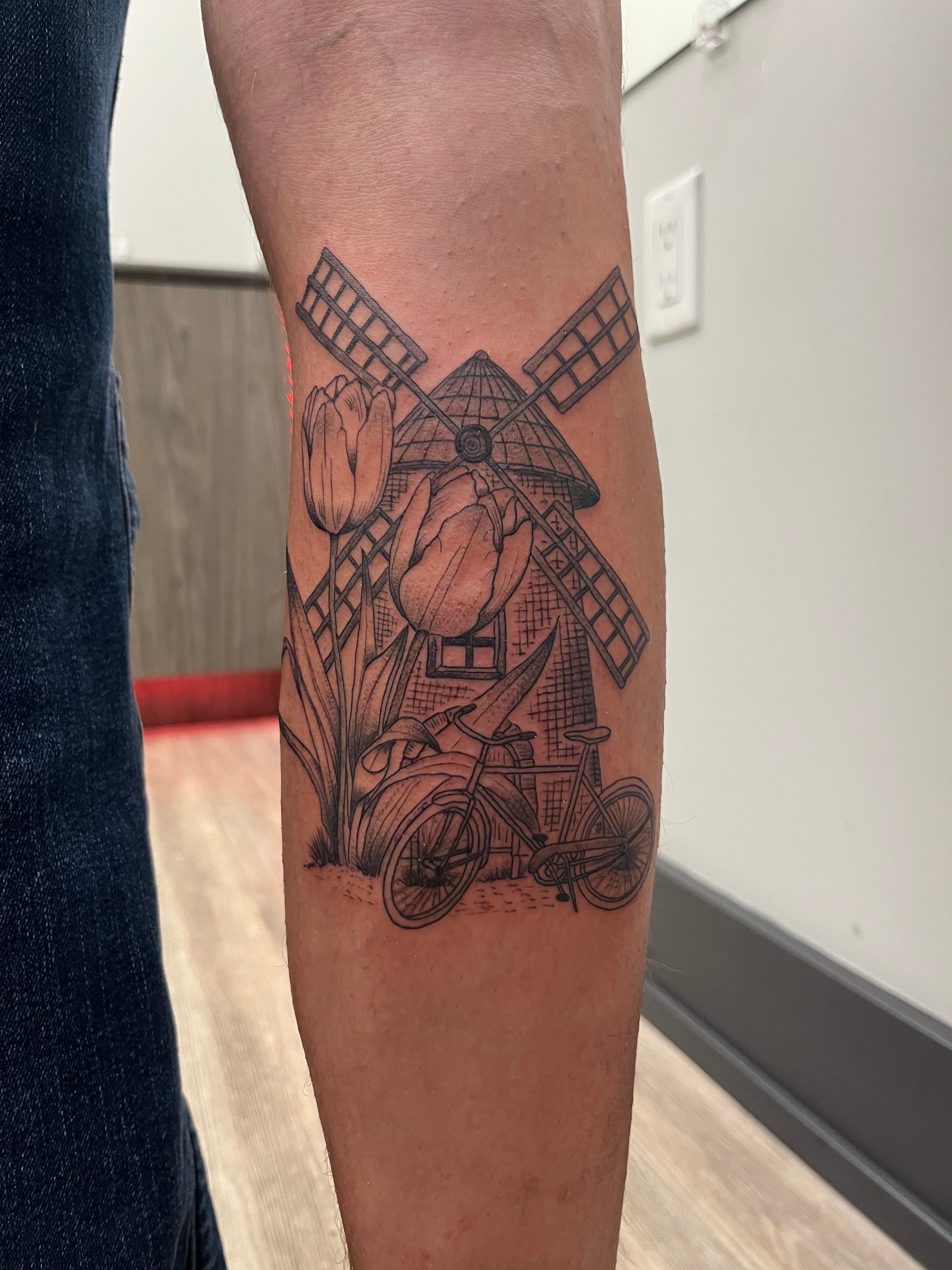 Black-and-grey fine-line windmill, tulips and bicycles tattooed on a forearm