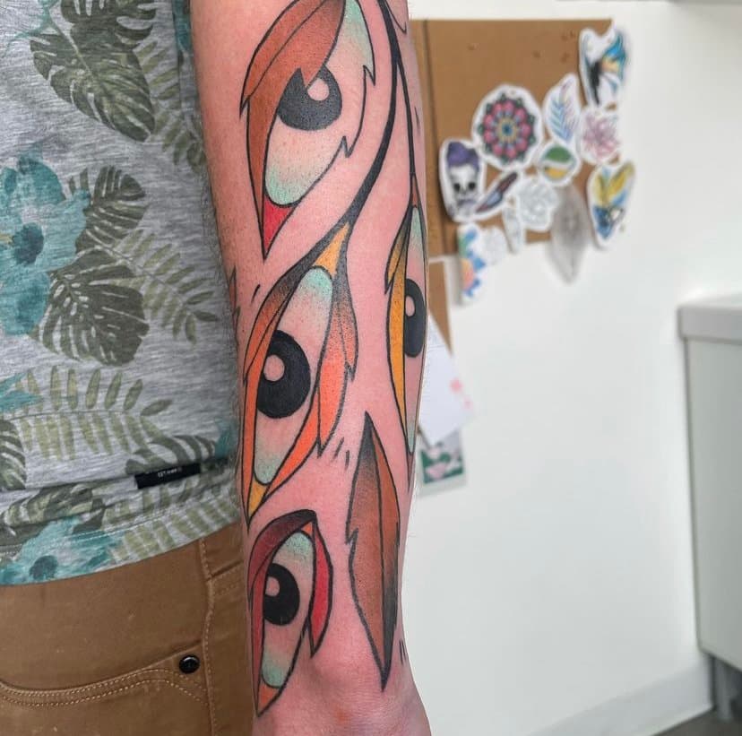Close-up of a colorful tattoo of large anime-style eyes on a person's arm, with a blurred background showing a bulletin board with character stickers.