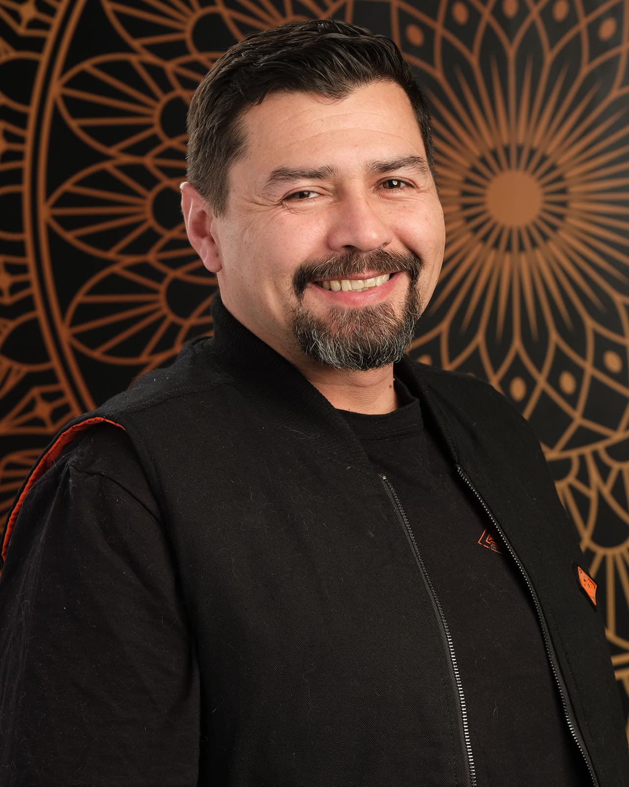 First-party studio portrait of Mario Mantilla in a black tee and black zip vest against the shop's black and orange mandala backdrop