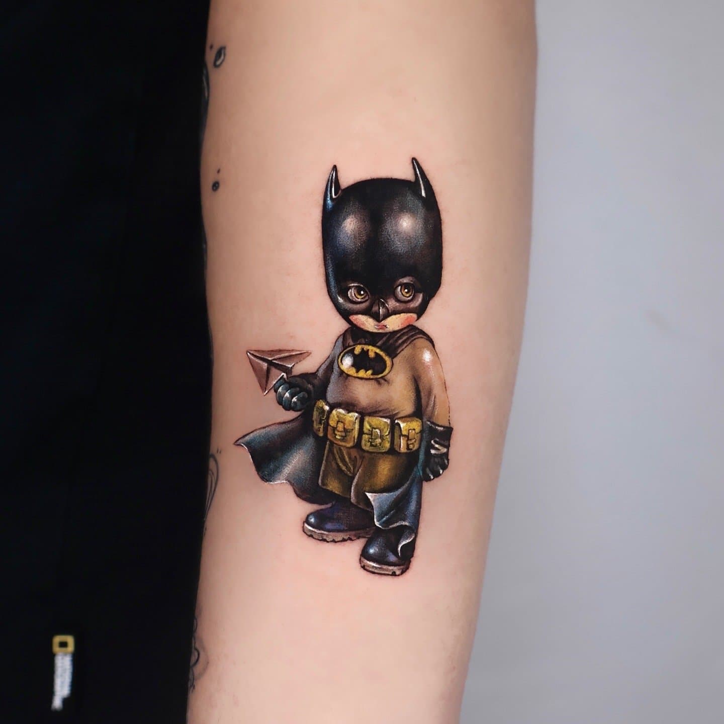 Tattoo of Batman with a child's face wearing a costume, holding a paper airplane.