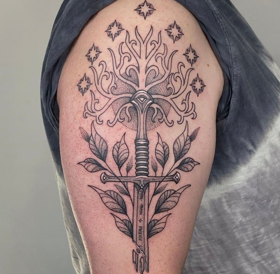 Black-and-grey upper-arm tattoo combining a fantasy sword, a stylized tree, leaves, and stars