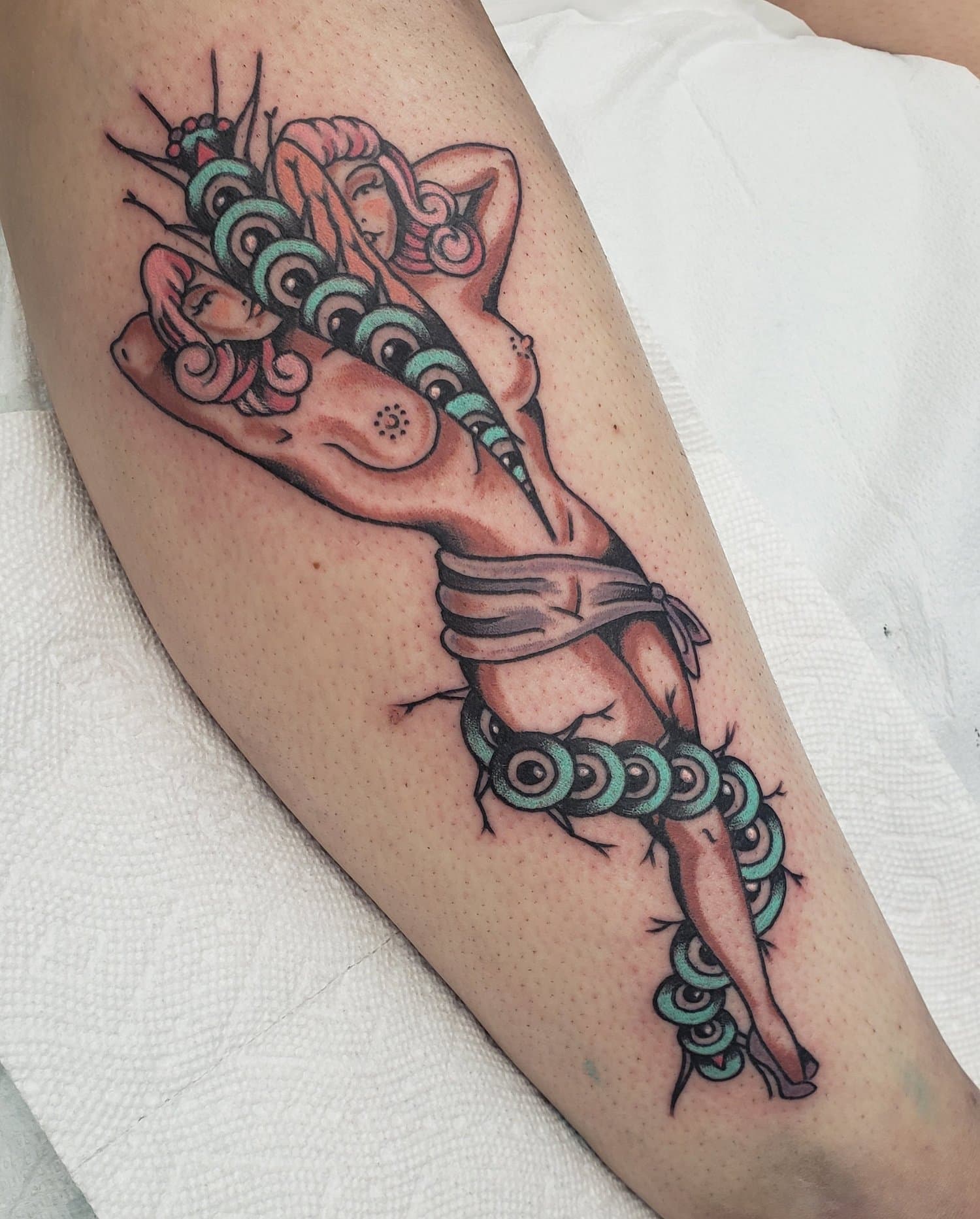 Tattoo of a nude woman with long hair, holding her head with one arm. Her body is wrapped with a line of green and black circular elements resembling a serpent