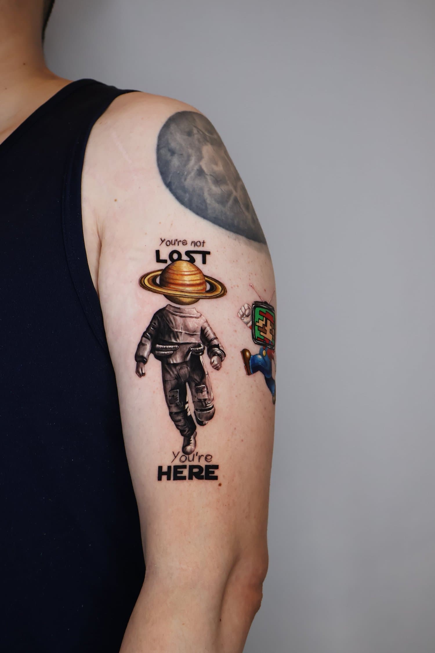 Tattoo of an astronaut with a planet on their head, text reads 'You're not lost, you're here', and a small cartoon character with a TV head nearby.
