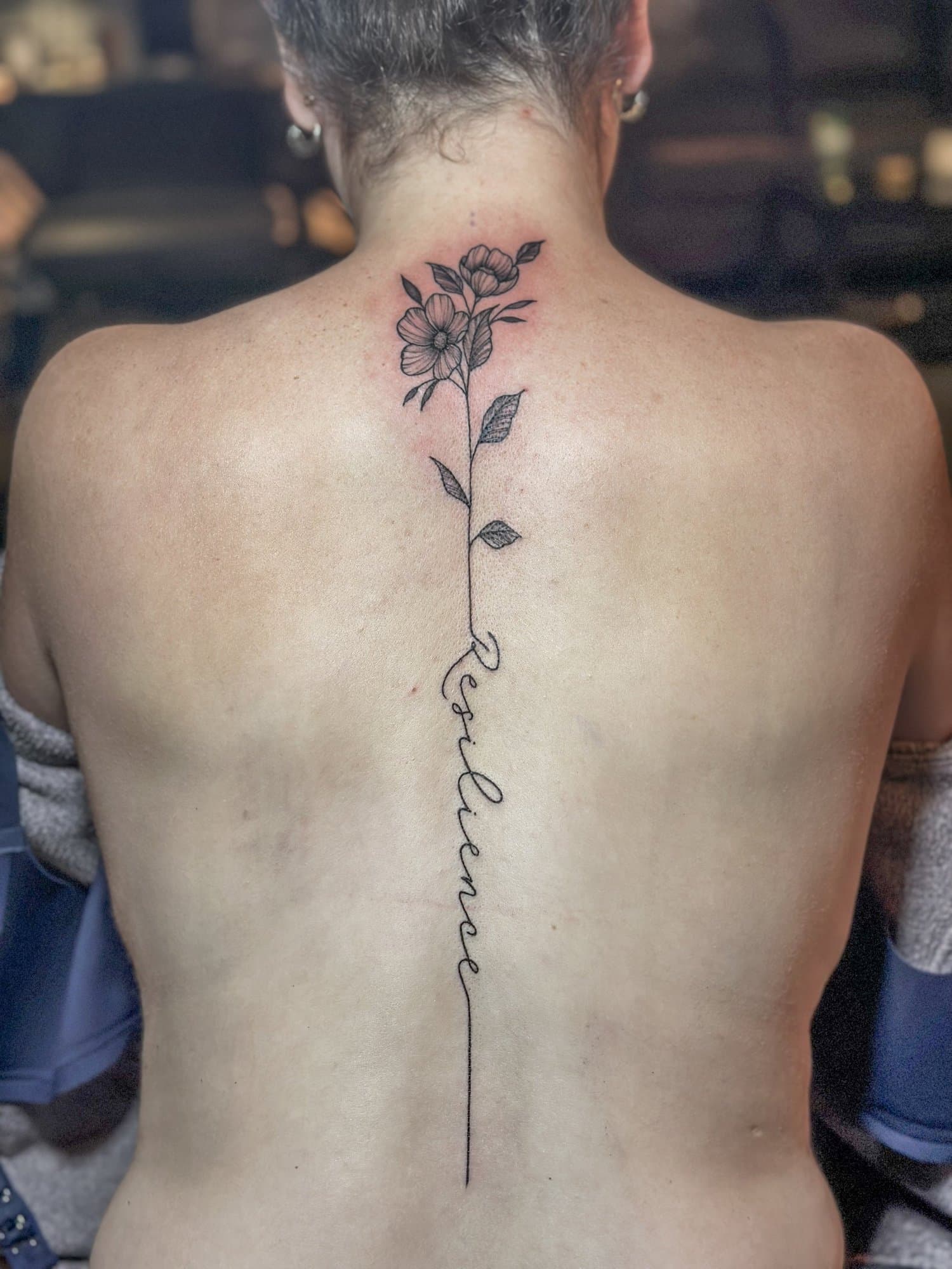 A person's back with a tattoo of a vertical line of flowers and leaves, with cursive writing running down the line.