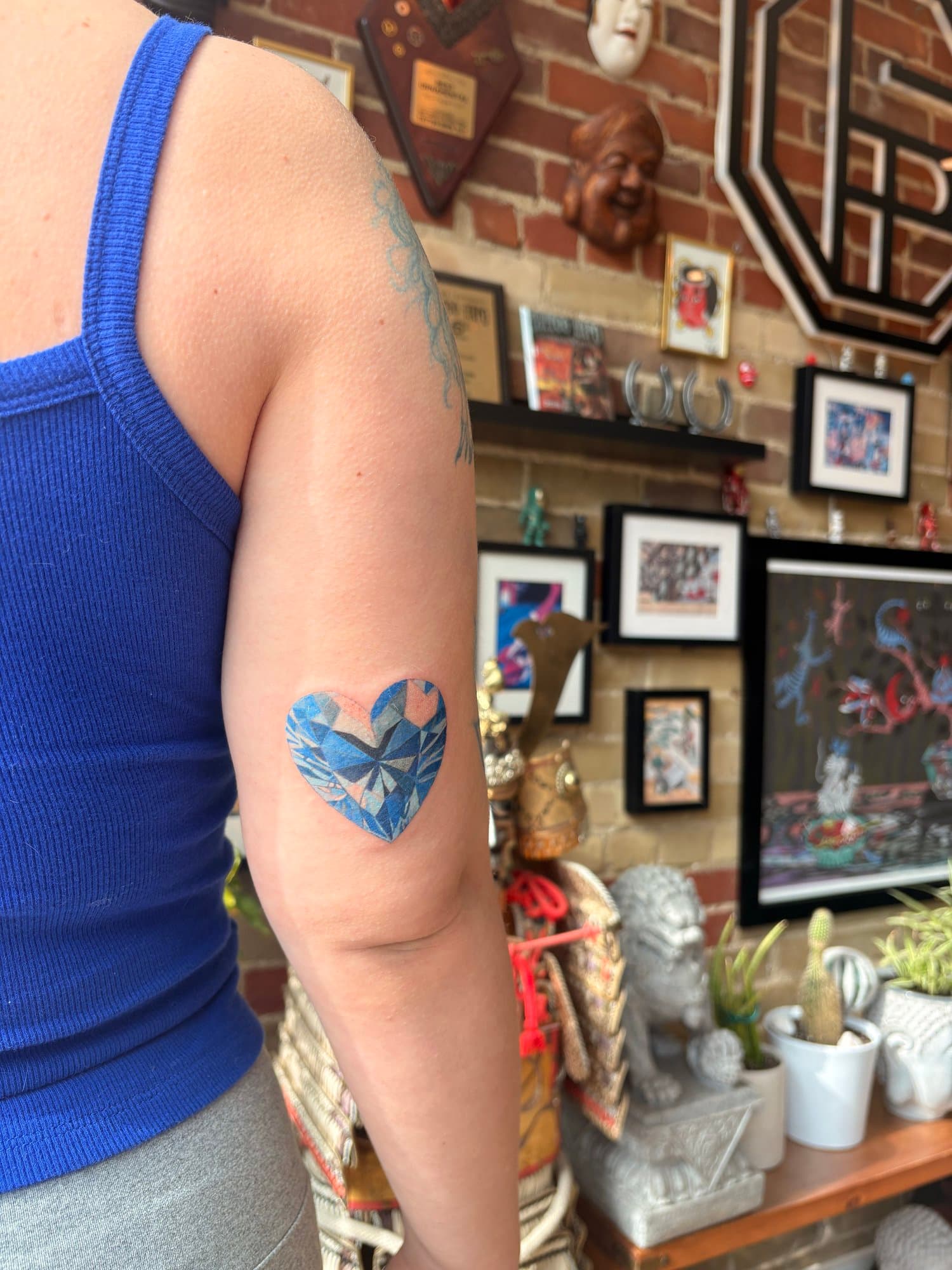 Close-up of a person's shoulder and arm, showing a heart-shaped tattoo, in front of a decorated wall with framed pictures and various decorative items.
