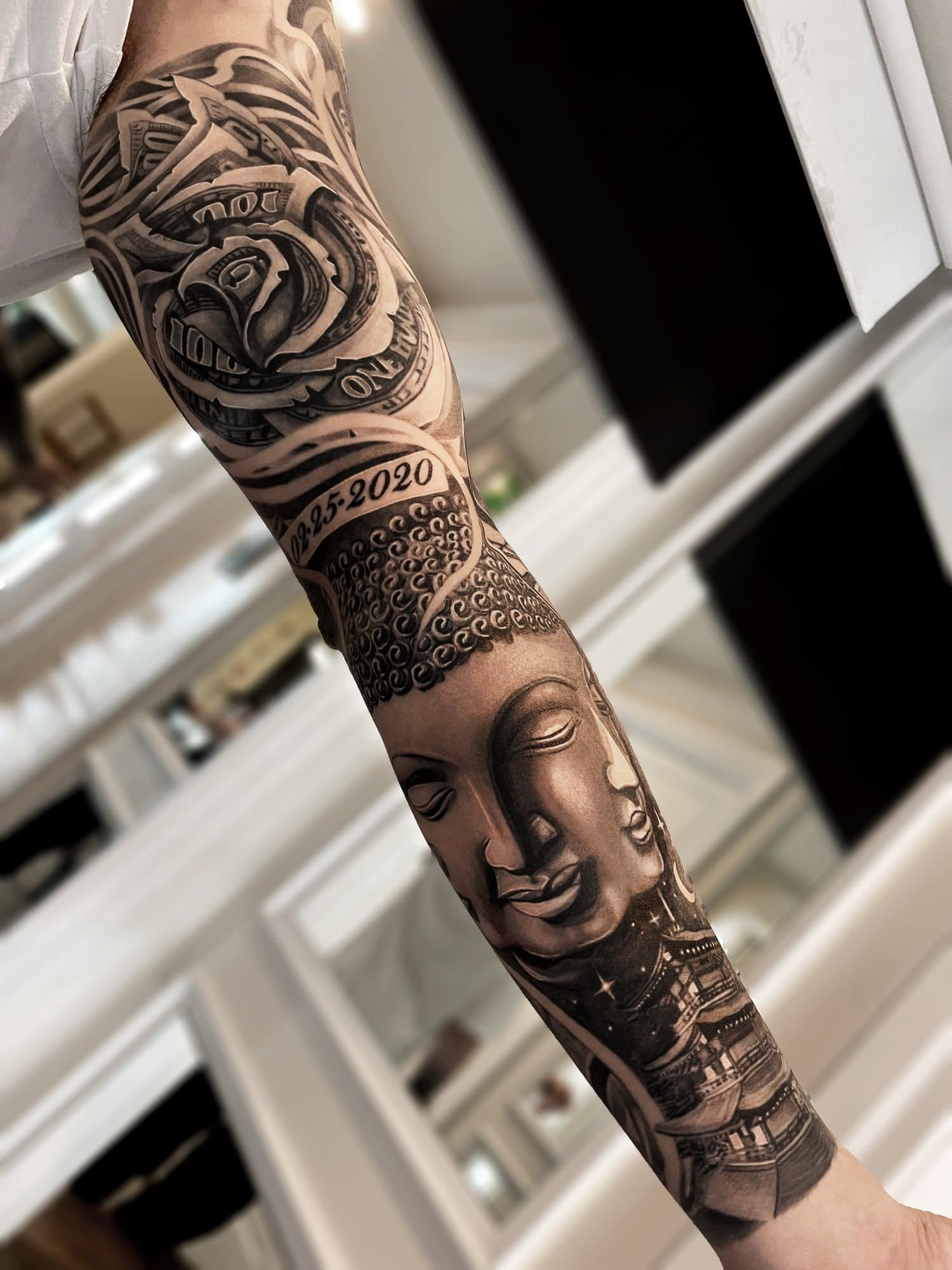 A person's arm tattoo featuring a face of a Buddha, a detailed eye, and various decorative elements in black and gray ink.