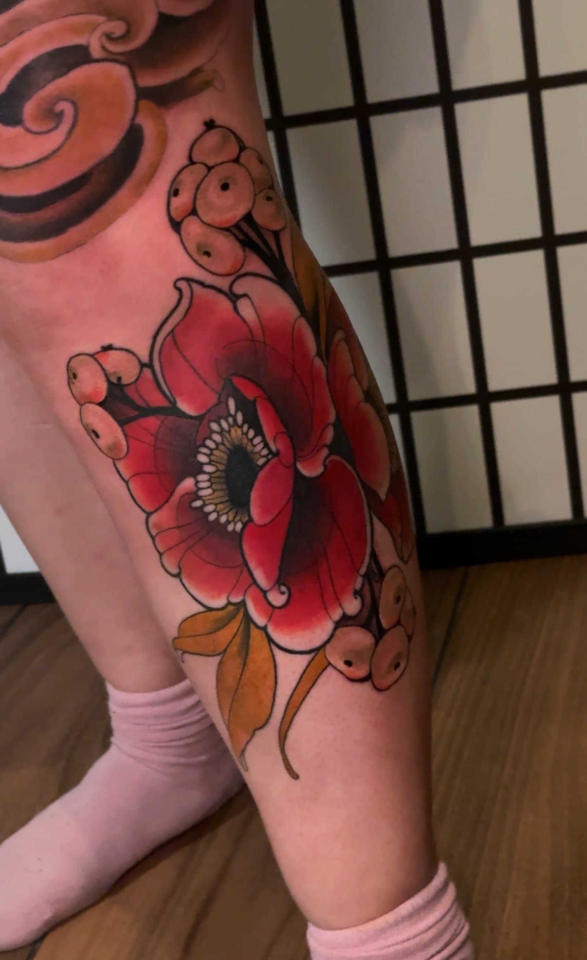 Tatouage d'une grande fleur rouge sur la cuisse, entourée de feuilles et de petites billes, avec un fond de porte-fenêtre en papier de riz.