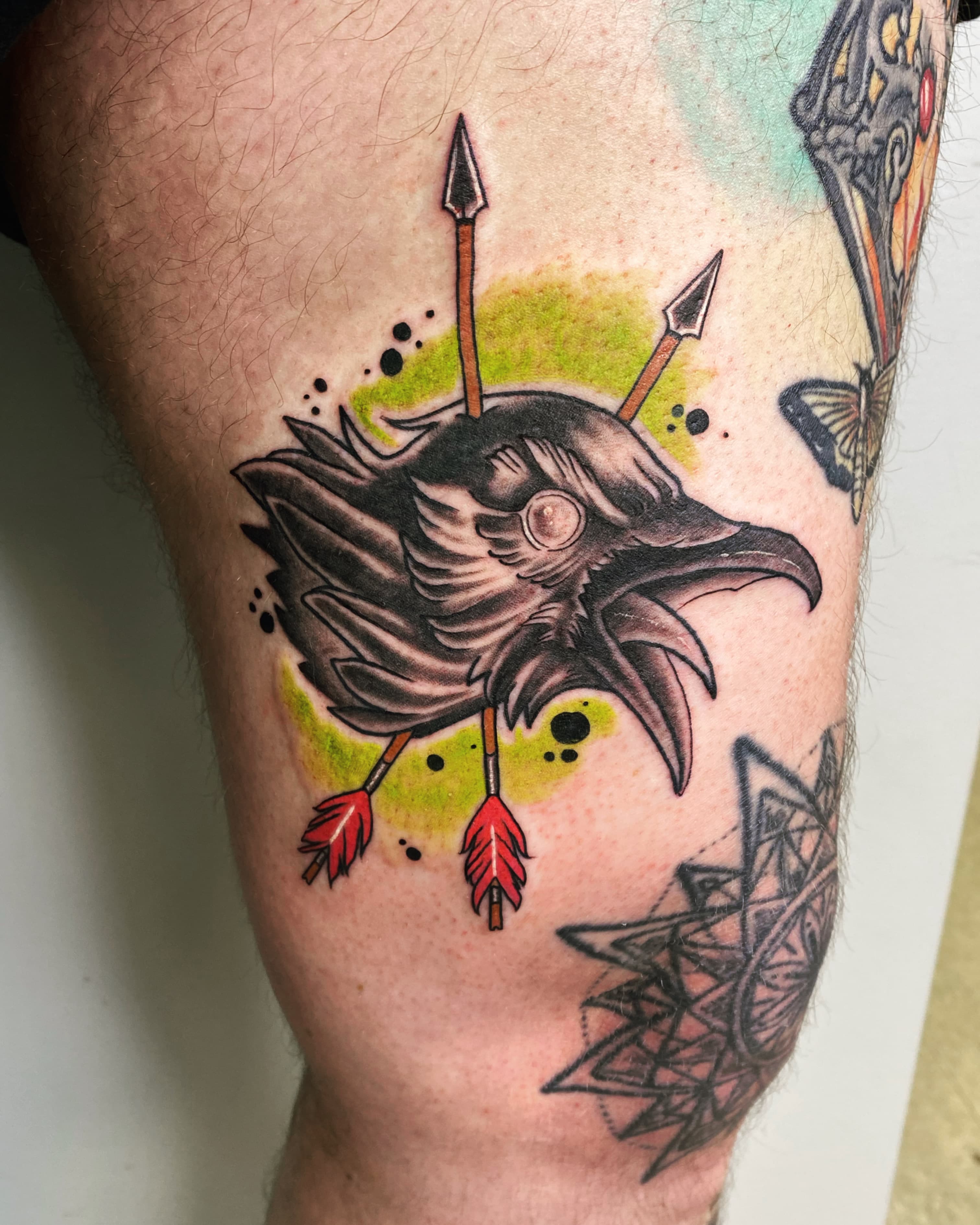 Neo-traditional leg tattoo of a raven head pierced by two arrows over a yellow-green wash, beside a geometric ornamental panel