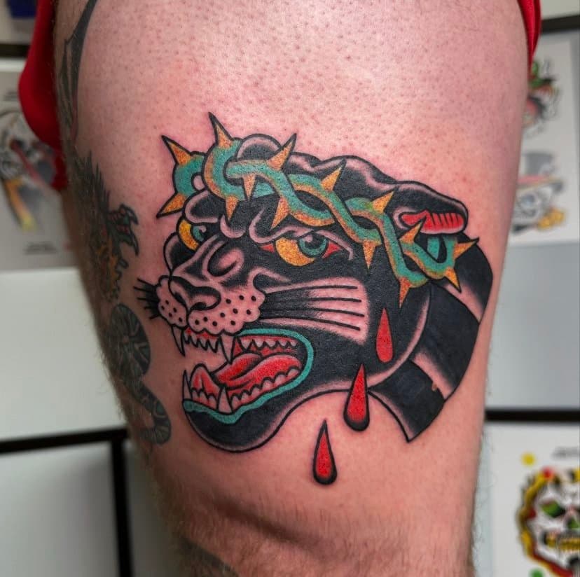 Tattoo of a fierce black panther with an open mouth showing sharp teeth, with red drops of blood and a spiked crown or headpiece, in a traditional tattoo style.